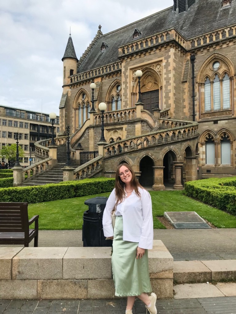 A young woman wearing a light green skirt and white blouse stands in front of a historic building in Scotland. She is smiling and wears glasses.